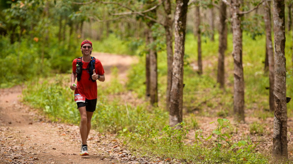 A male trail runner in a red shirt, black shorts, and a hydration backpack jogs along a dirt path surrounded by lush greenery and tall trees. He wears a red headband and sunglasses, giving a thumbs-up while holding a small water bottle. The setting is a forested area with a mix of green foliage and fallen leaves on the path, suggesting a warm, outdoor adventure.