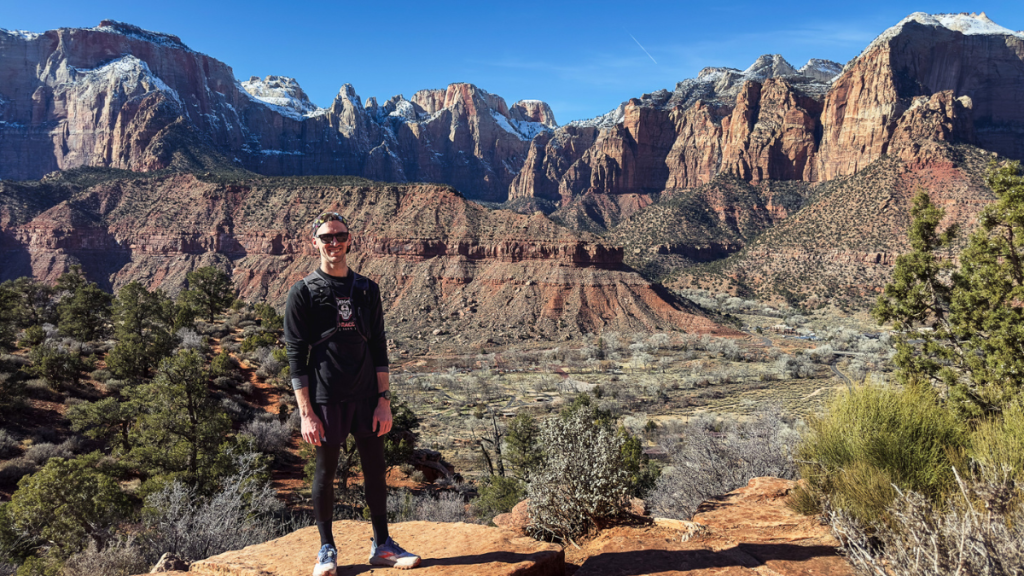 A man wearing sunglasses, a black long-sleeve shirt, black athletic pants, and white sneakers stands on a rocky overlook with a smile. Behind him, a vast desert landscape with towering red rock formations and snow-capped peaks stretches under a clear blue sky. The scene appears to be in a national park, with rugged terrain, scattered trees, and deep canyons creating a breathtaking backdrop.