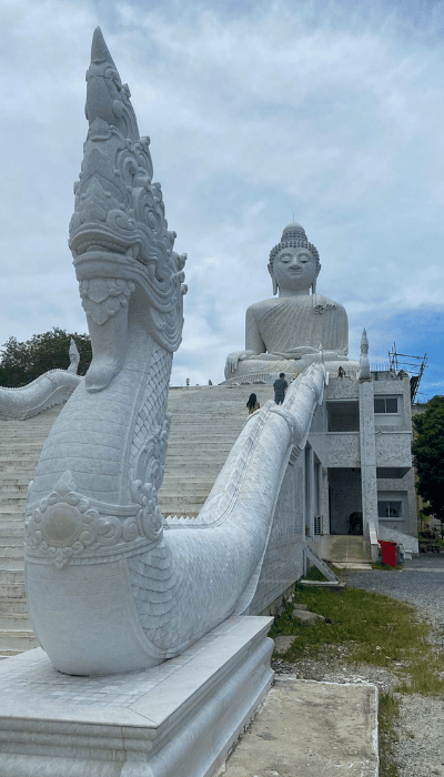 A wide staircase guarded by ornate white naga sculptures leads to Phuket’s towering Big Buddha statue under a cloudy blue sky.
