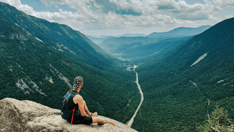A hiker sits on a rocky ledge overlooking a dramatic, forested mountain pass with a road winding through the valley below, capturing the panoramic view from the summit of the Mount Willard Trail.