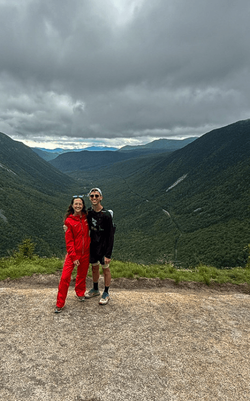 Two runners smile at the camera at the summit of Mount Willard, with a dramatic view of Crawford Notch stretching behind them. The lush green valley contrasts with the moody overcast sky, capturing the scenic reward at the end of the Mount Willard Trail.