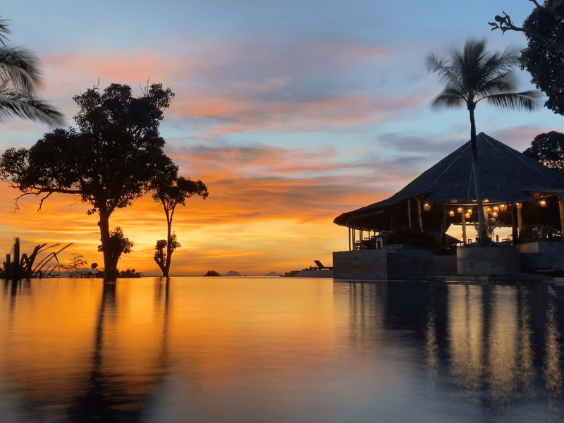 An infinity pool reflects vibrant orange and blue hues as the sun sets behind silhouetted palm trees and a glowing open-air restaurant on Koh Yao Yai, a bit more off the beaten path in Phuket.