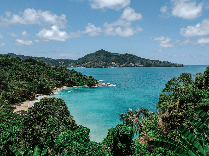 A turquoise bay framed by dense tropical forest and a small sandy beach, with forested hills and calm waters stretching into the distance.