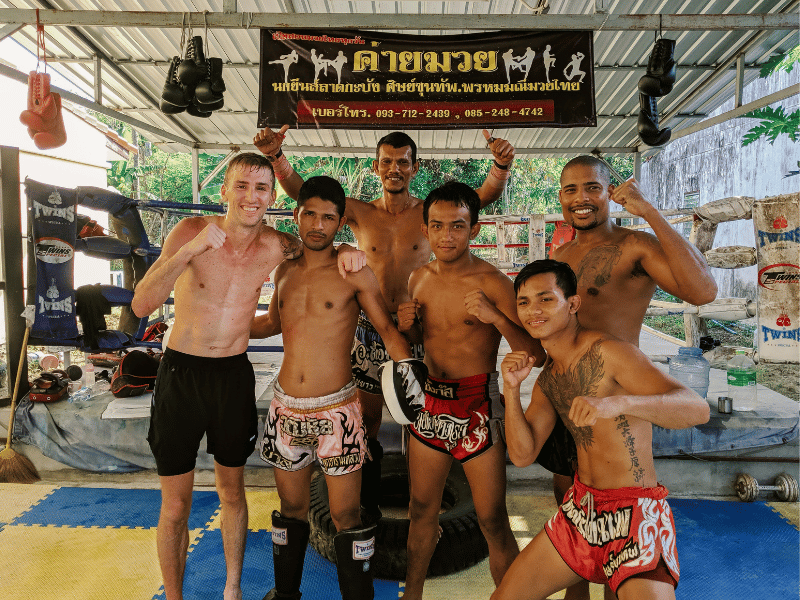 A group of men in Muay Thai shorts pose with smiles and fighting stances after training, standing on padded mats under a metal-roof gym with Thai signage overhead.