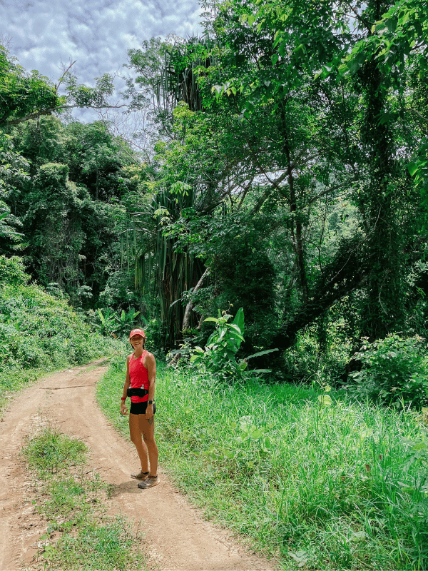 A woman in athletic gear stands on a dirt trail winding through lush jungle, surrounded by towering greenery and tropical plants.