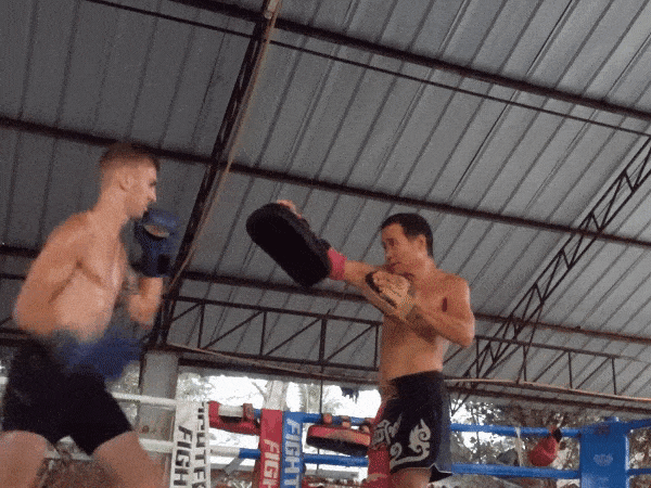 A shirtless fighter in black shorts throws a fast punch at a coach holding pads during a training session inside a boxing ring.