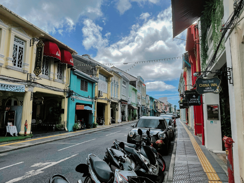 A quiet street in Phuket Old Town lined with pastel-colored Sino-Portuguese buildings, red awnings, parked scooters, and signs for local cafés and massage shops under a partly cloudy sky.