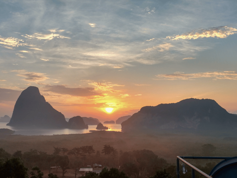 The sun rises over misty limestone cliffs and calm water, casting a golden glow on the dramatic karst landscape viewed from a peaceful overlook — a highlight for those seeking non touristy things to do in Phuket.