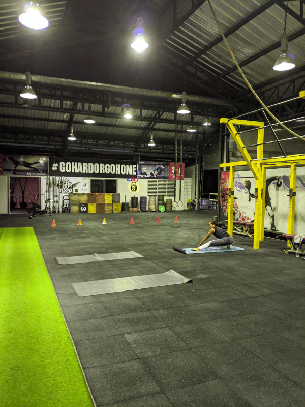 A lone woman stretches on a yoga mat inside a large industrial gym with rubber flooring, colorful cones, wall art, and a bold sign reading “#GOHARDORGOHOME.”