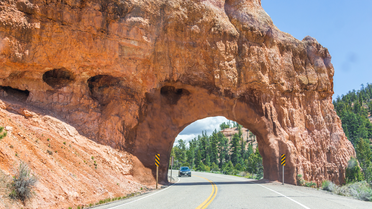 A paved road winds through a natural archway carved in the red rock formations of Red Canyon, with a car driving through the tunnel.