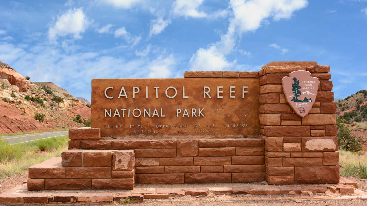 The Capitol Reef National Park sign, made of red rock with the park name in raised lettering, stands in front of a blue sky with white clouds and a scenic road.