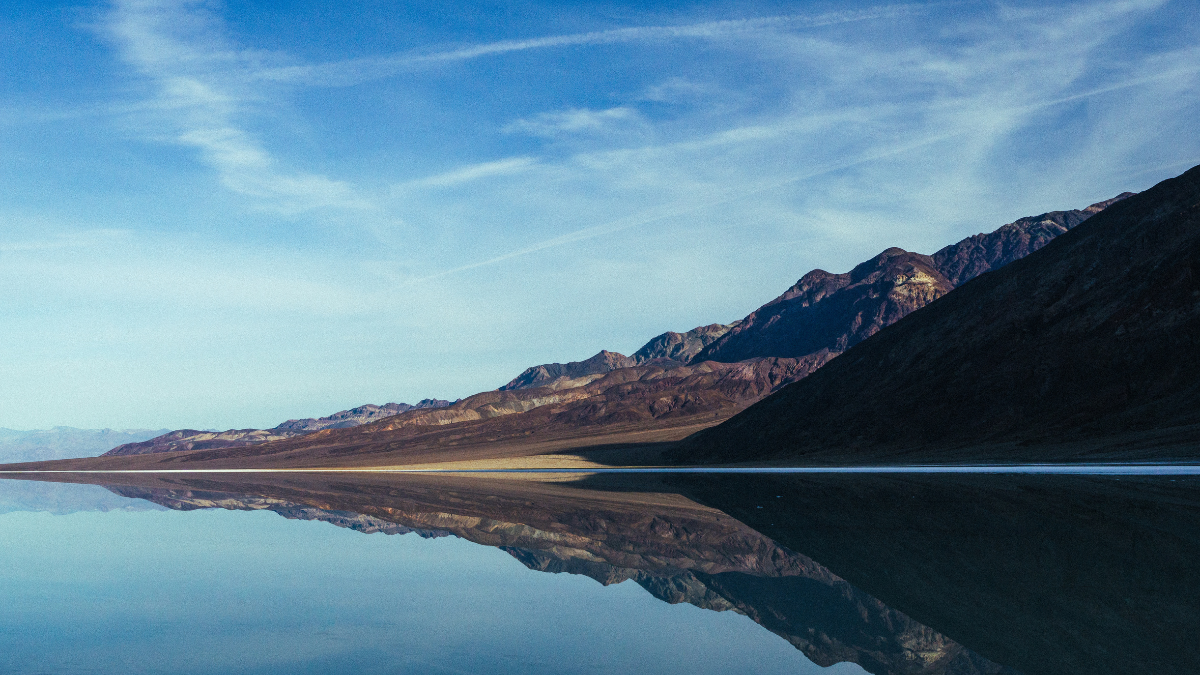 erene landscape of Death Valley with calm water reflecting the rugged mountains and blue sky with wispy clouds.