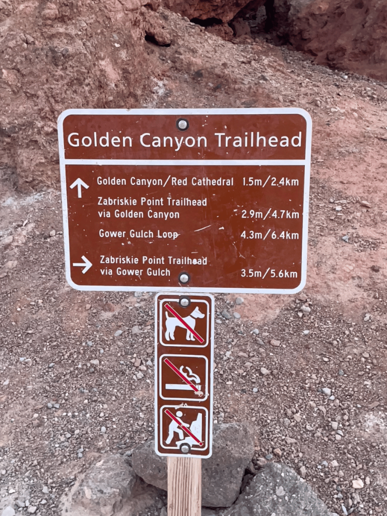 Trailhead sign for Golden Canyon/Gower Gulch in Death Valley National Park, displaying directions and distances to various points, including Golden Canyon/Red Cathedral, Zabriskie Point via Golden Canyon, and Gower Gulch Loop. Below, prohibitory signs indicate no dogs, no smoking, and no vehicles are allowed on the trails.