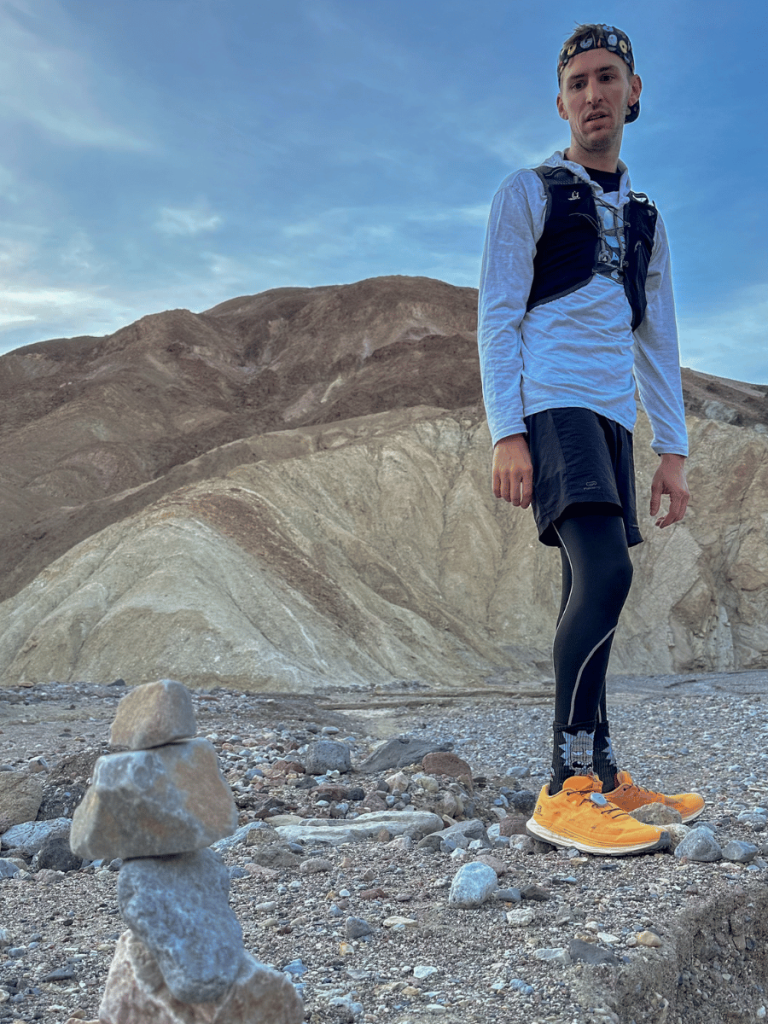 A hiker in a white long-sleeve shirt, black shorts, and bright orange shoes stands on a rocky trail in Gower Gulch, Death Valley. In the foreground, a small cairn of stones is neatly stacked, with the barren, folded hills of the gulch in the background under a pale blue sky.