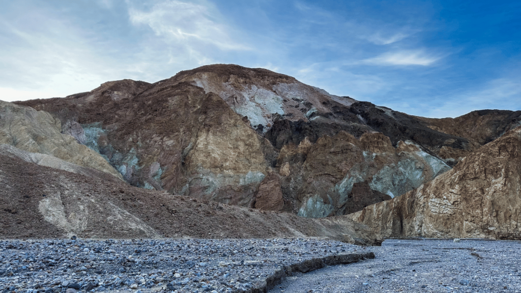 Variegated mineral deposits create a striking palette on the hills near Zabriskie Point in Death Valley, with a foreground of a gravelly wash and a clear blue sky above.
