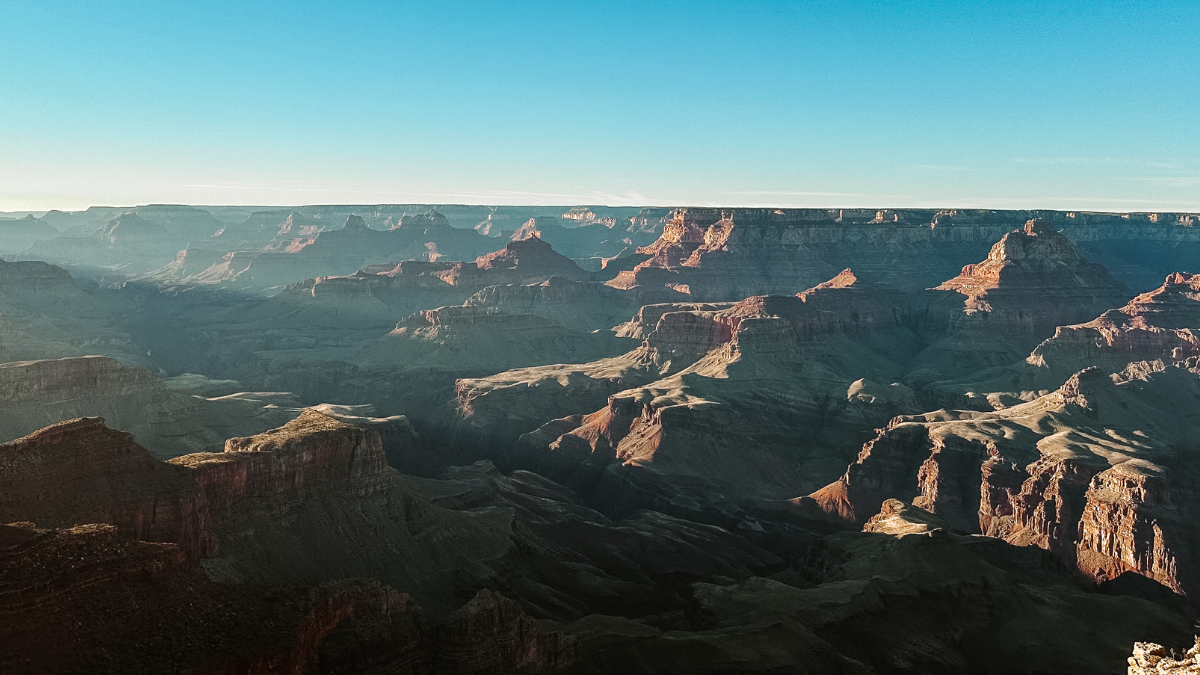 Vast panorama of the Grand Canyon at sunrise, with layered rock formations in shades of red and brown stretching to the horizon.