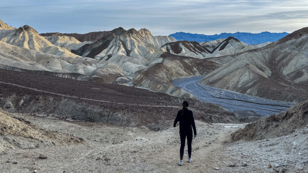A lone hiker in black attire walks down a rugged trail at Gower Gulch in Death Valley, with a spectacular view of multicolored hills and distant mountains under a soft blue sky.