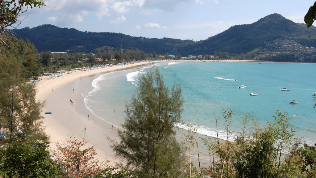 The calm shores of Kamala Beach in Phuket, with soft waves, golden sands, and a scenic view of the surrounding hills.
