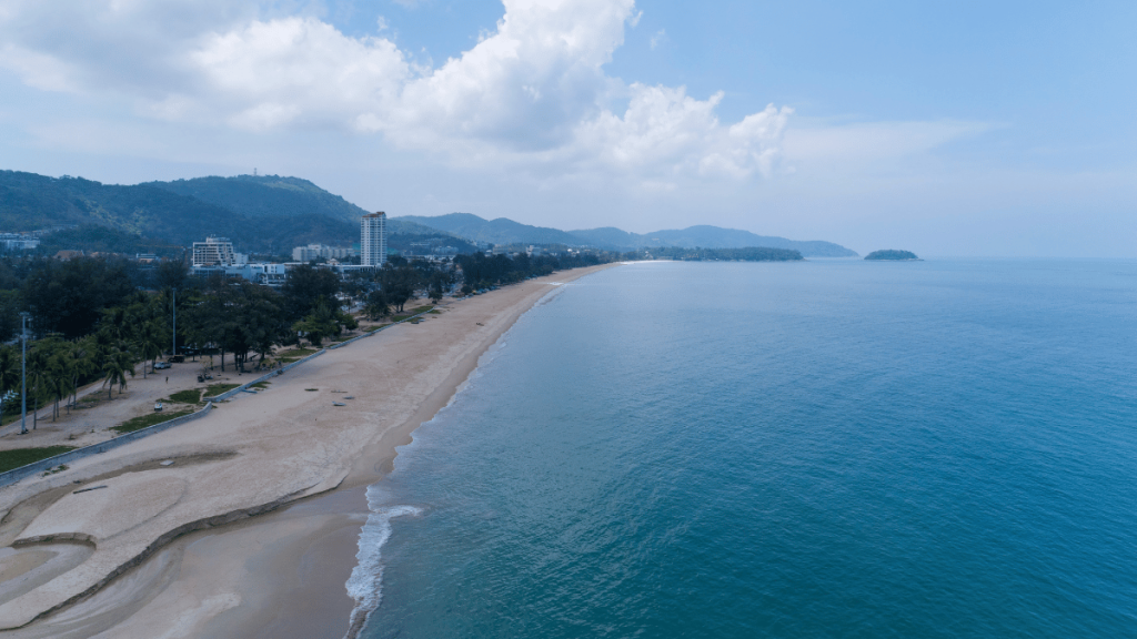 A wide aerial shot of Karon Beach in Phuket, showing the long stretch of sandy beach, azure waters, and the expansive seaside town.