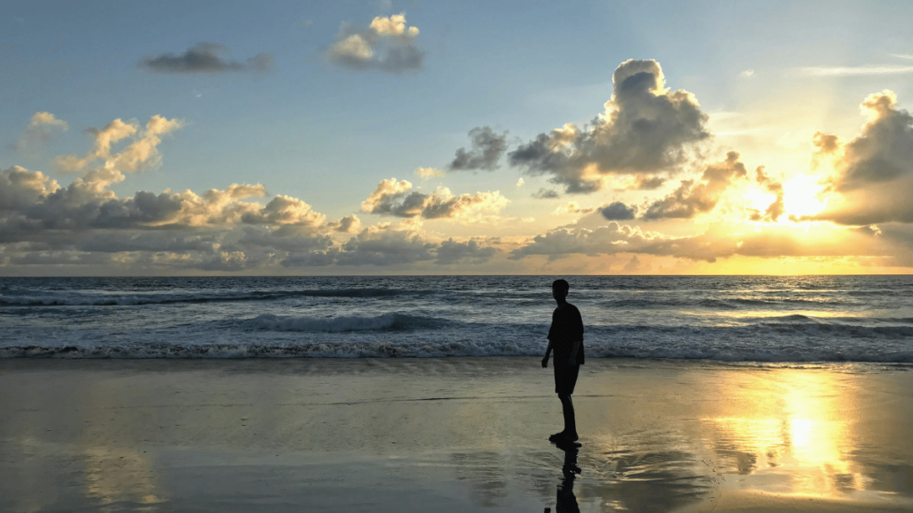 A solitary figure stands on Karon Beach in Phuket, silhouetted against the golden sunset reflecting off the calm ocean waters.