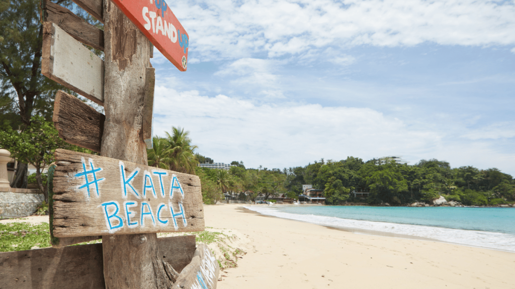 Hand-painted sign reading '# Kata Beach' with a blue hashtag symbol, with the beautiful sandy shores of Kata Beach in the background." Nai Harn Beach: "A peaceful view of Nai Harn Beach in Phuket, featuring a hammock tied between two trees, with the beach's clear turquoise waters and sandy shores in the distance.