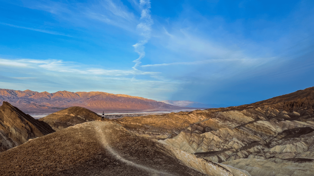 A hiker stands at a viewpoint on the ridge of the Golden Canyon Trail, overlooking the expansive Death Valley terrain with Manly Beacon in the distance, under a vast blue sky with wispy clouds.