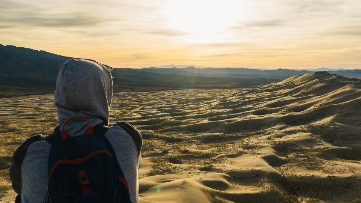 A person wearing a backpack and hoodie gazes at the sun setting over rolling sand dunes.
