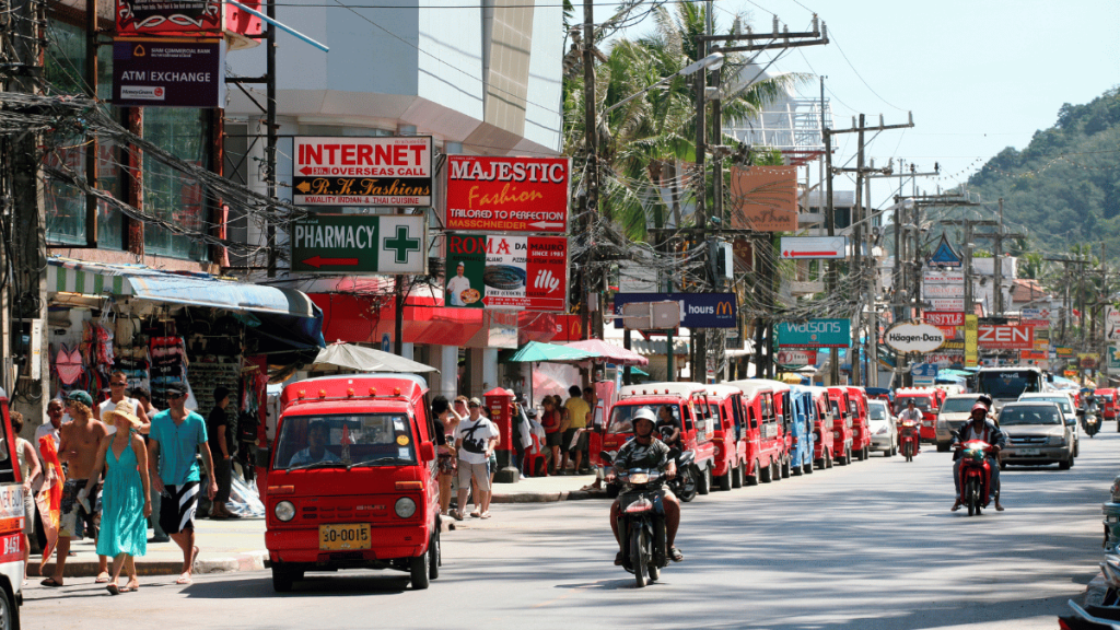 Busy street scene at Patong Beach in Phuket, lined with tuk-tuks and local shops, bustling with tourists and locals under a clear blue sky.