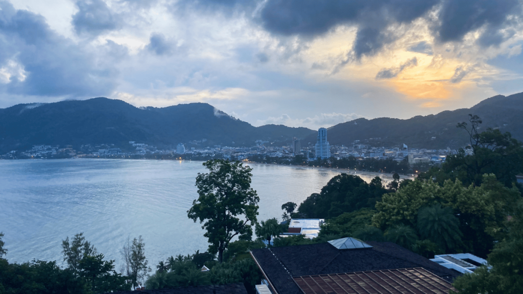 The sun sets over Patong Beach in Phuket, casting a golden glow over the calm sea and silhouetted onlookers on the shore.