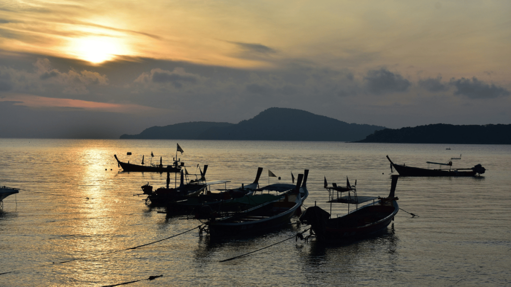 Silhouettes of traditional longtail boats anchored in the calm waters of Rawai at sunset, with the golden sun descending behind a mountainous horizon in Phuket.