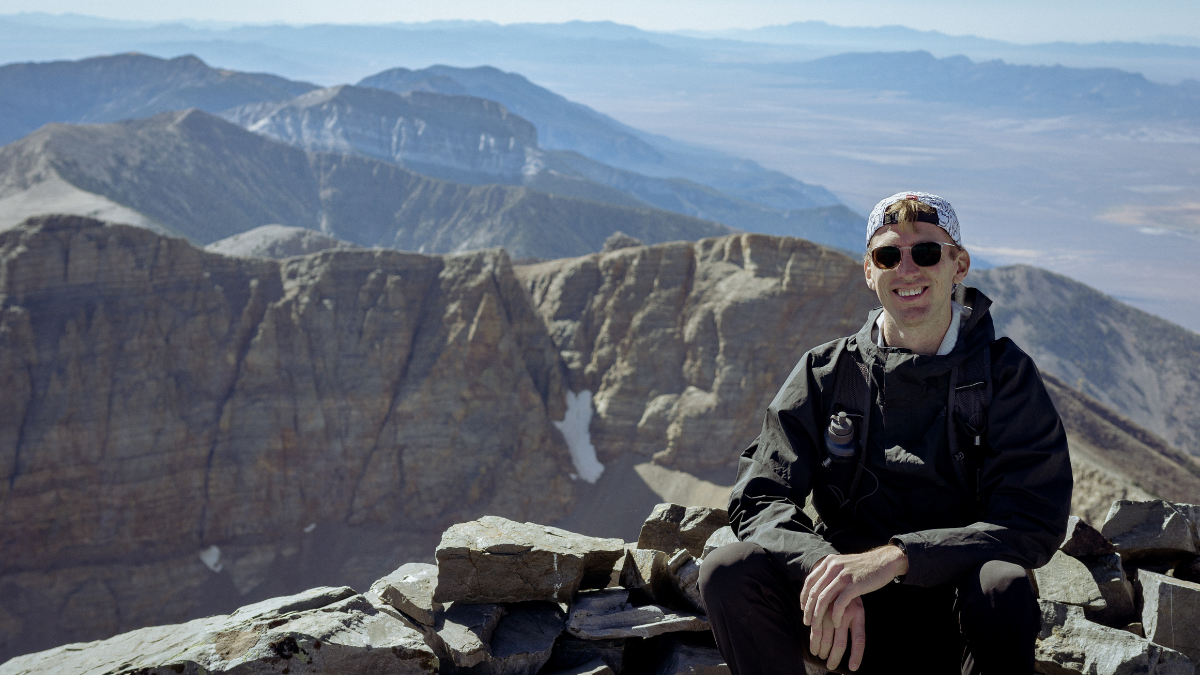 A man sits at the top of Wheeler Peak with giant mountains in the background