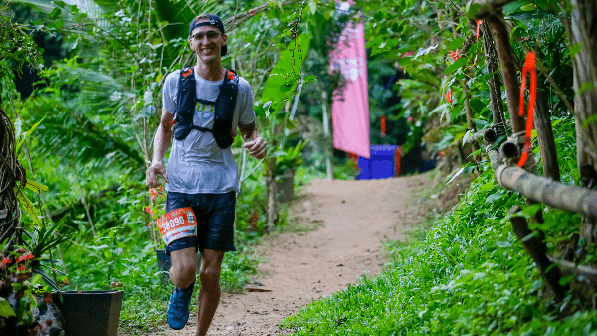 A trail runner is smiling as he runs through a thick green forest on a dirt trail while wearing a white shirt and gray shorts