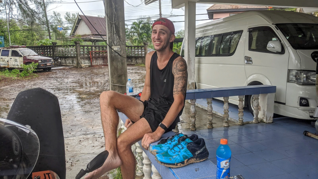 A smiling man in a black tank top and shorts sits on a blue-tiled porch, removing his muddy shoes after what appears to be a run in wet conditions. He wears a red backward cap and has a tattoo on his left arm. A pair of blue trail running shoes and a bottle of orange sports drink rest on the ledge beside him, while a white van and a rain-soaked yard with parked vehicles are visible in the background.