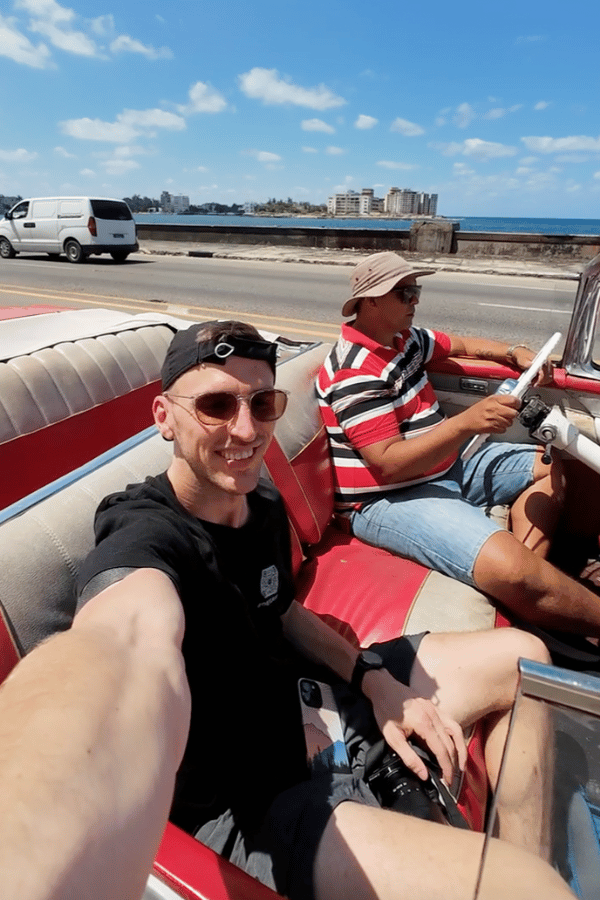 A tourist takes a selfie while riding in the back seat of a red and white vintage convertible along Havana’s Malecón, with a Cuban driver at the wheel. The ocean and city skyline stretch out in the background under a bright, sunny sky.