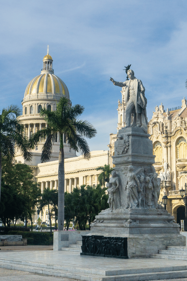 A marble statue of Cuban national hero José Martí stands with an outstretched arm in front of the El Capitolio building in Havana, Cuba. The dome of the Capitolio glows in the sunlight behind swaying palm trees, with ornate architecture from the Gran Teatro de La Habana partially visible on the right.