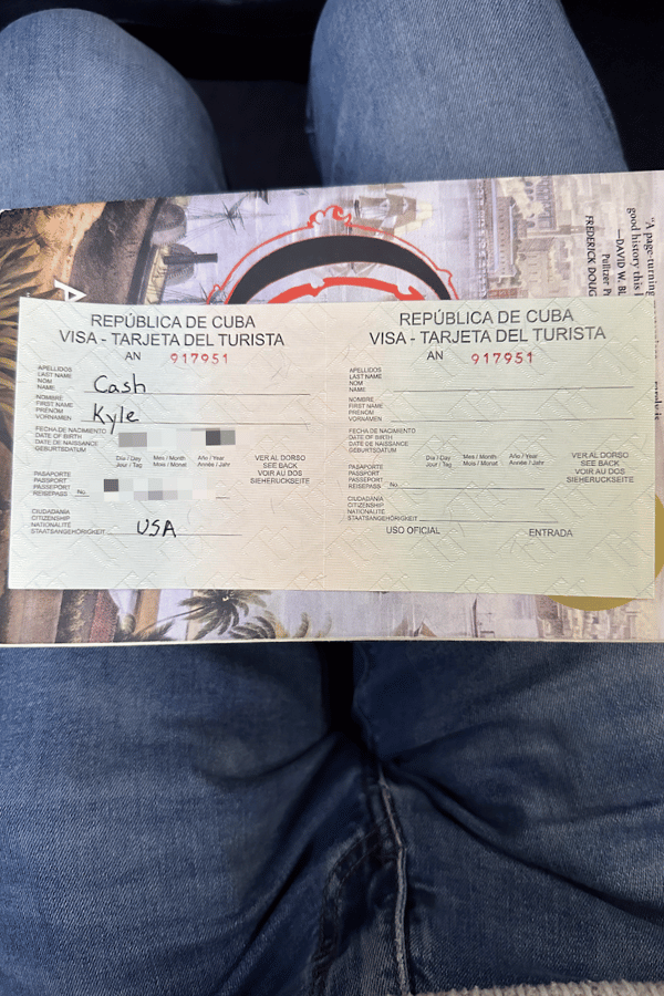 A person holds an official Cuban tourist visa card open across their lap, with handwritten name and nationality visible on the left side. The background features jeans-clad legs and a partially visible travel-themed design behind the visa form.