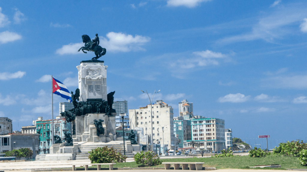 Statue of Máximo Gómez on horseback stands atop a tall white marble monument near the Malecón in Havana, Cuba. The monument is surrounded by sculpted figures and a Cuban flag waves nearby, with colorful buildings and the sea in the background.