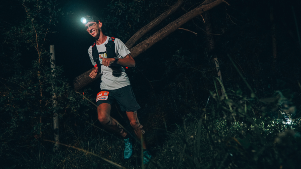 A male trail runner wearing a headlamp and a hydration pack runs through a dense forest at night. He is dressed in a light-colored shirt, dark shorts, and bright blue trail shoes, with a race bib attached to his shorts. The glow of his headlamp illuminates the dark surroundings, casting shadows on the trees and highlighting the rugged terrain.
