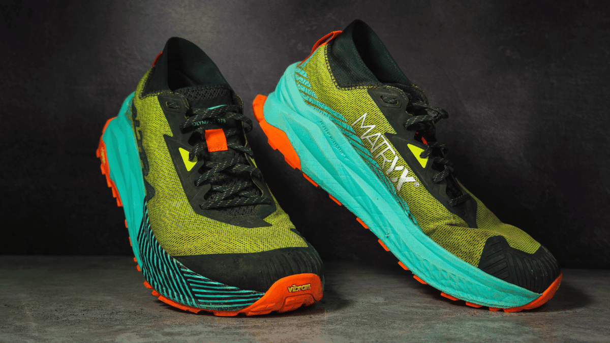 A pair of Altra Olympus 2.75 trail running shoes is displayed against a dark backdrop, showcasing their bold design with olive green uppers, turquoise midsoles, and Vibram outsoles in bright orange. The MATRYX and Olympus 2.75 branding emphasize the shoes’ durability and cushioning, making them standout pieces of trail running gear.