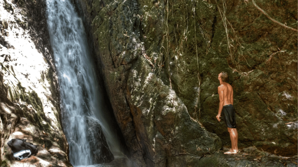 A shirtless man stands at the base of a waterfall, gazing up at the cascading water on the Bang Pae to Tonsai trail in Phuket, Thailand. The waterfall flows down a steep, rocky cliff surrounded by lush greenery, creating a tranquil and refreshing natural scene. A small backpack lies on the ground nearby, suggesting the man is taking a break to enjoy the serene environment.