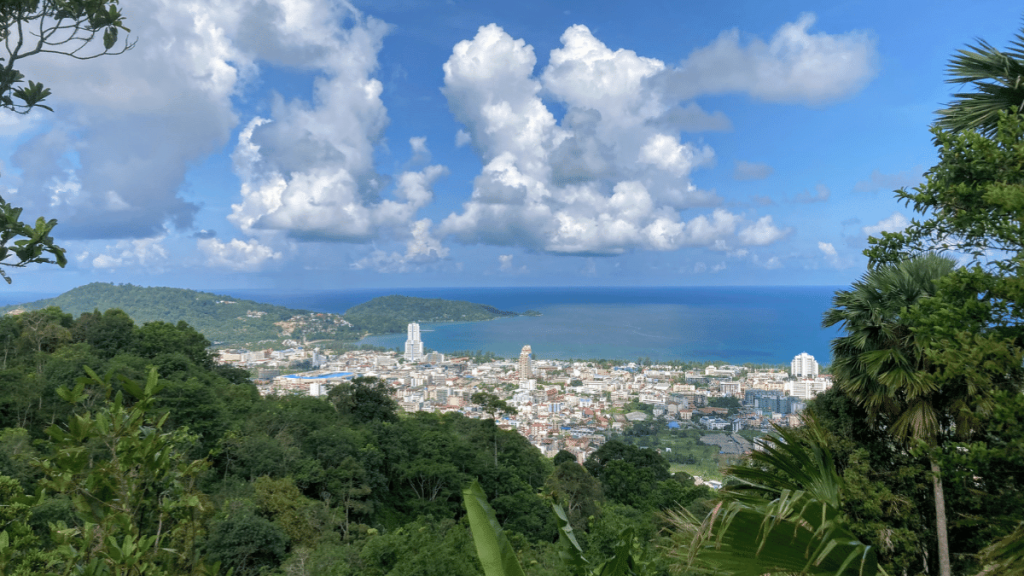 A panoramic view of Patong, Phuket, Thailand, taken from a high vantage point. The bustling city is surrounded by lush green hills and overlooks the clear blue waters of the Andaman Sea. The scene is framed by tropical vegetation, and the sky is filled with fluffy white clouds, creating a picturesque and vibrant landscape.