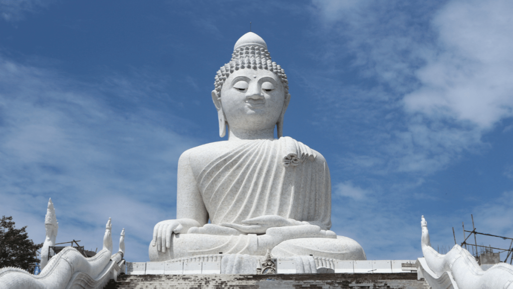 A large, white marble statue of the Big Buddha sits majestically against a bright blue sky in Phuket, Thailand. The statue features intricate details, with serene facial expressions and flowing robes, embodying peace and spirituality. The surrounding area includes decorative elements and construction scaffolding, indicating ongoing work to maintain or enhance the site.