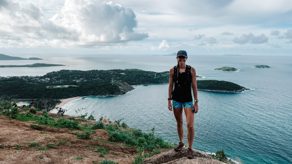 A person in athletic gear, including a cap and shorts, stands on a rocky outcrop at Black Rock Viewpoint in Phuket, Thailand. They are smiling, with a scenic view of the coastline, lush green hills, and small islands in the background. The sky is partly cloudy, enhancing the breathtaking landscape.