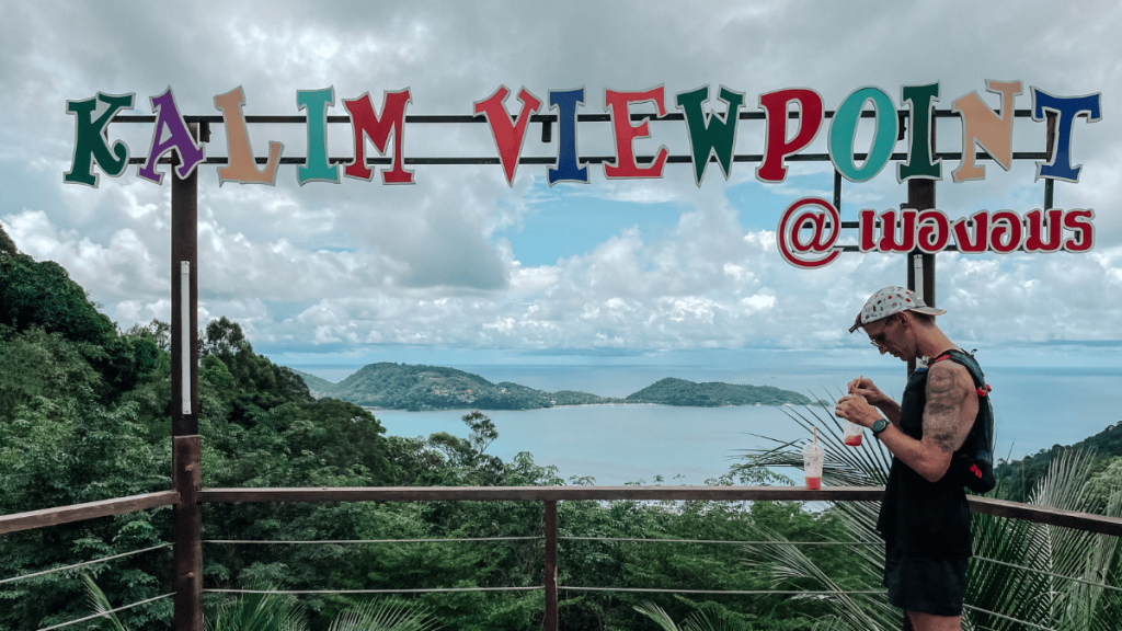 A person wearing a cap and backpack is seen standing on a viewing platform at Kalim Viewpoint in Phuket, Thailand. The colorful sign "Kalim Viewpoint" is prominently displayed above, with lush green hills and the ocean visible in the background. The person appears to be adjusting their gear or using a device.