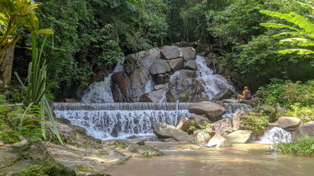 A person with a backpack sits near the cascading waters of Kathu Waterfall in Phuket, Thailand. The waterfall flows over large rocks surrounded by dense green foliage, creating a serene and picturesque natural scene. Sunlight filters through the trees, adding a peaceful ambiance to the location.