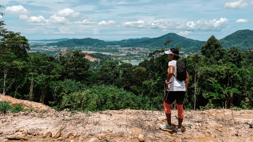 A runner wearing a cap, backpack, and athletic gear stands on a dirt trail overlooking a scenic valley from Manik Mining Trail in Phuket, Thailand. The lush green forest spreads out below, with distant hills and a small town visible under a partly cloudy sky. The view suggests a moment of rest and appreciation of the natural beauty and expansive landscape.