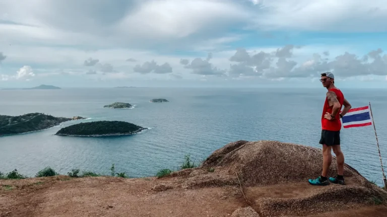 Man in athletic gear stands on a rocky cliff next to a Thai flag, overlooking the ocean and nearby islands under a cloudy sky. The scene captures a scenic viewpoint in Phuket highlighting both the physical challenge and the stunning coastal views.