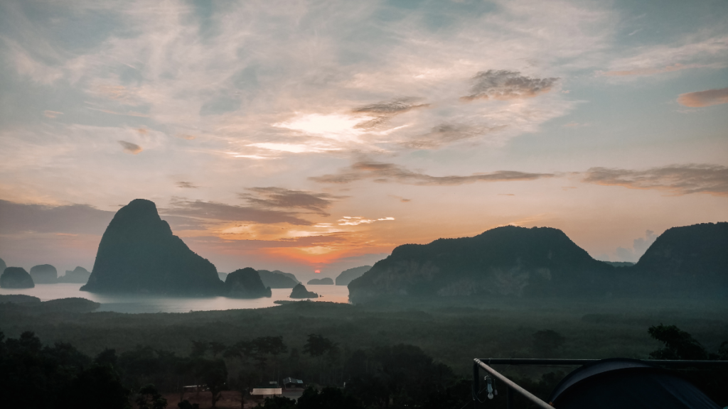 A breathtaking sunrise view from Samet Nangshe Viewpoint in Phang Nga, Thailand. The scene features dramatic limestone karsts rising from the bay, with the sky painted in hues of orange, pink, and purple. The mist-covered landscape and calm waters add a serene and mystical quality to the panoramic vista.