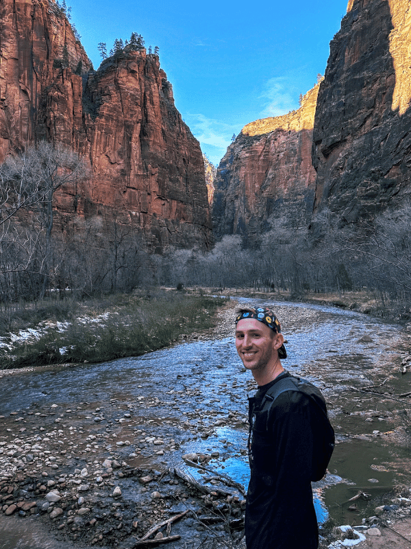A hiker smiles by a shallow, rocky river with steep red rock cliffs towering on either side under a bright blue sky in Zion National Park.