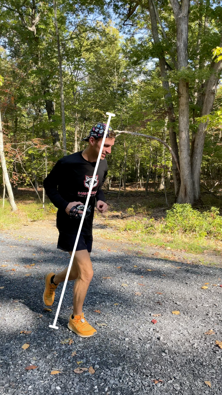 A man is running uphill on a gravel path in a forest, leaning forward. He is wearing a black long-sleeve shirt, black shorts, orange shoes, and a patterned cap. A white line is running through the center of his body vertically showing a slight forward lean to run uphill faster.
